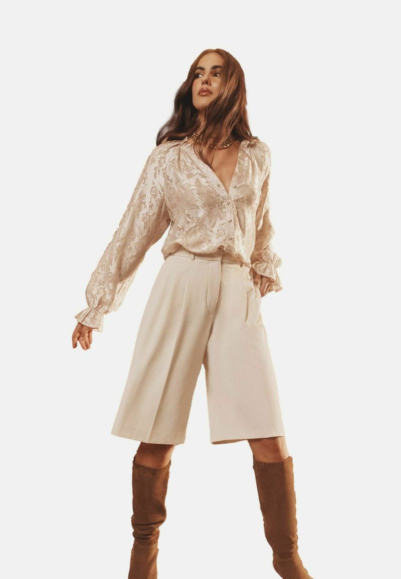Hellbeige Bluse mit floralem Muster, V-Ausschnitt und langen, gerafften Ärmeln, kombiniert mit cremefarbenen, weiten Hosen und knielangen, braunen Wildlederstiefeln.