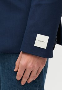 Χέρι που ακουμπάει πάνω σε μπλε τζιν παντελόνι, φορώντας μπλε σκούρο σακάκι με λευκή ετικέτα Calvin Klein στο μανίκι κοντά στον καρπό.