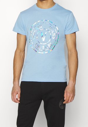 Homme portant une chemise à manches courtes bleu clair avec le logo argenté brillant "Versace Jeans Couture" et un pantalon noir avec un petit logo assorti.