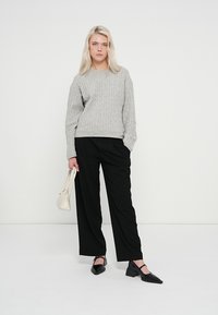 Szürke kötött pulóver, fekete bő fazonú nadrág, fekete hegyes orrú balerina cipő és egy kis fehér kézitáska. Egyszínű háttér.