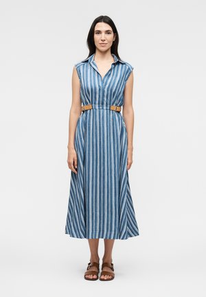 Kvinde står iført en ærmeløs blå og hvid vertikalt striped midi kjole med krave, brun bælte og brune sandaler på en ensfarvet baggrund.