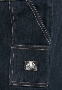 Dark denim fabric close-up featuring a patch pocket with yellow stitching and a square logo in black and white reading "SOUTHPole Original."