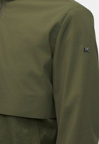 Manche de veste vert olive avec un motif texturé subtil en dessous et un petit logo argenté « MK » sur le haut du bras.