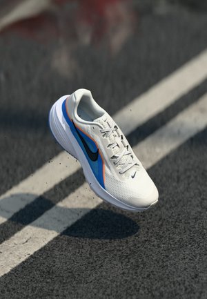 Scarpa da corsa bianca con accenti blu e arancioni e logo Nike nero, appare sospesa sopra una strada asfaltata con segnaletica bianca a linee.