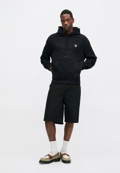 Haut à capuche noir avec poche avant et cordon de serrage, associé à un short noir et des chaussures à enfiler vertes et brunes. Le mannequin se tient devant un fond uni.