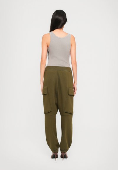 Woman with long dark hair stands facing away, wearing a gray sleeveless top, olive green cargo pants, and brown high-heeled shoes.