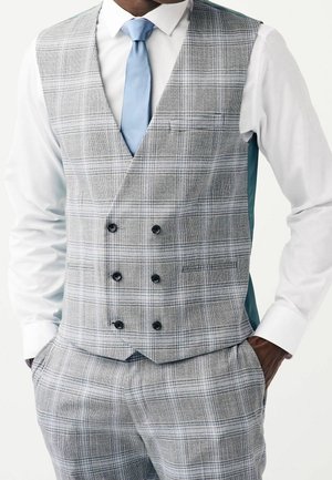 Homme portant un gilet croisé à carreaux gris et un pantalon assorti, chemise blanche et cravate bleu clair, mains dans les poches, sur fond blanc.