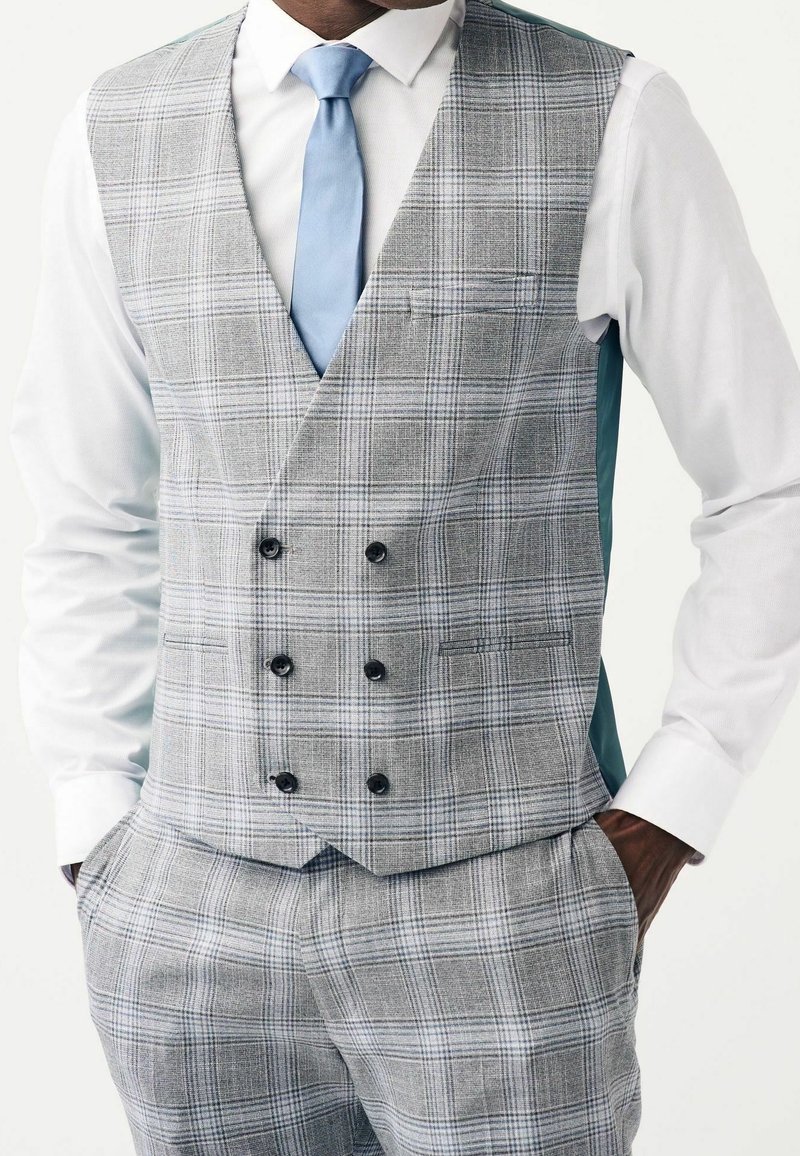 Homme portant un gilet croisé à carreaux gris et un pantalon assorti, chemise blanche et cravate bleu clair, mains dans les poches, sur fond blanc.