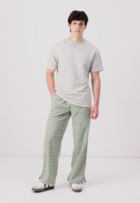 Un tricou gri cu mânecă scurtă și margini desfăcute, asortat cu pantaloni cu carouri în nuanțe de verde deschis. Modelul poartă adidași gri și albi. Fundal curat.