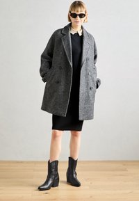 Šedý vlněný dvouřadový kabát s velkými knoflíky, nošený přes černé šaty. Obsahuje černé kožené kotníkové boty s blokovým podpatkem.