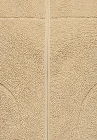 Beige fleece-stoff med en sentral sølvglidelås og buede sømdetaljer på en myk, teksturert overflate.