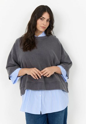 Vrouw met lang donker haar, die een grijze oversized korte mouw top draagt, gelaagd over een blauw-wit gestreept overhemd en blauwe spijkerbroek.