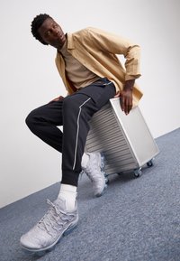Szare sportowe buty z przezroczystą podeszwą, czarne spodnie z białymi bocznymi paskami oraz beżowa koszula noszona na beżowym topie. Srebrna walizka ustawiona obok.