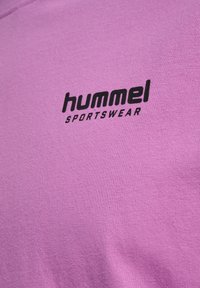 Közeli kép lila szövetről, fekete "hummel SPORTSWEAR" logóval, amely a felületen van nyomtatva.