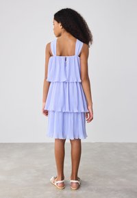 Robe violette claire plissée avec des couches superposées, des bretelles larges et une fermeture éclair au dos. Le tissu a une texture douce et un tomber fluide.