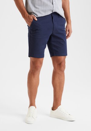 Dunkelblaue Shorts aus Baumwollstoff, im klassischen Schnitt mit seitlichen Taschen, getragen mit weißen Sneakers und einem hellgrauen Hemd mit Knopfleiste.