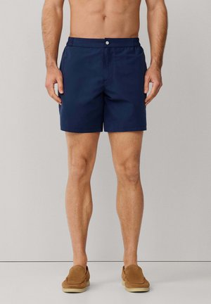 Mężczyzna ubrany w granatowe, dopasowane szorty i jasnobrązowe buty wsuwane, stojący na jednolitym tle.