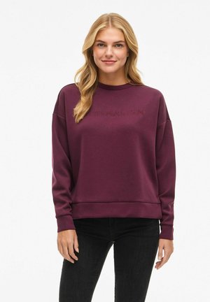 Blonde woman wearing a long-sleeve burgundy sweatshirt with "MINIMALISM" embossed on the chest and black pants, smiling.