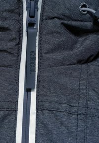 Tmavě šedá bunda s texturovanou látkou, která má výrazný zip s logem na poutku a reflexní detaily na přední straně.