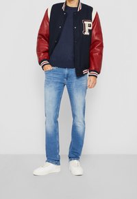 Námornická varsity bunda s červenými koženými rukávy, bílými pletenými manžetami a nášivkou 'P'; v kombinaci se světle modrými džíny a bílými teniskami.