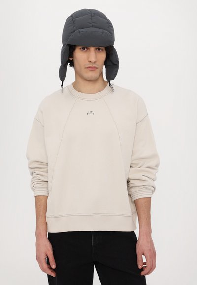 Sweatshirt beige clair avec des poignets côtelés, arborant un petit logo sur la poitrine. Associé à un bonnet à lacets noir foncé.