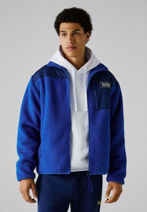Chaqueta de forro polar azul con cremallera frontal, superficie texturizada, acentos en el hombro en color marino y un bolsillo en el pecho que presenta el logo de "Red Bull Racing". Sudadera debajo.