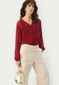 Blusa a fiori rossi con scollatura a V, maniche lunghe e texture morbida. Abbinata a pantaloni beige a vita alta e una borsa dorata.
