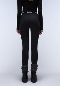 Czarne legginsy z wysokim pasem z detalem w postaci logo. Modelka nosi grube, szare, włókninowe ochraniacze na nogi oraz czarne botki na platformie.