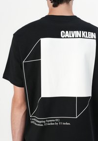 Juodas medvilninis marškinėliai su balta grafika, kuri vaizduoja persidengiančius kvadratus gale, viršuje esant užrašui "CALVIN KLEIN".