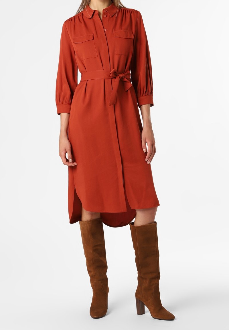 Robe-chemise couleur rouille avec deux poches poitrine, manches longues, taille ceinturée et ourlet asymétrique. Associée à des bottes montantes marron jusqu'aux genoux.