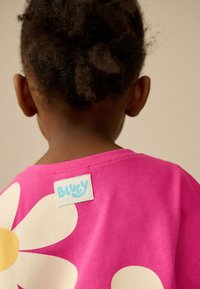 Maglietta rosa acceso con grande fiore bianco e centro giallo. Vista posteriore mostra un'etichetta "BLUEY" cucita sul collo. Tessuto morbido in cotone.