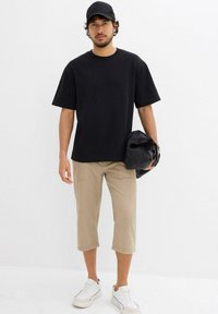 T-shirt nera a maniche corte, pantaloni corti beige, sneakers bianche, in mano una giacca nera. Design semplice, tessuto leggero, vestibilità rilassata.