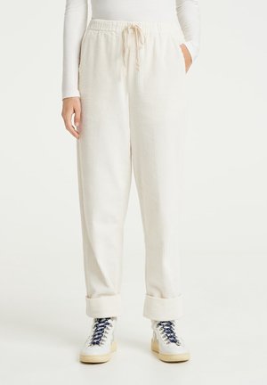 Person iført hvide ribstrikkede bukser med elastisk talje og snor, manchetterne er rullet op, kombineret med hvide sneakers med mørke snørebånd.