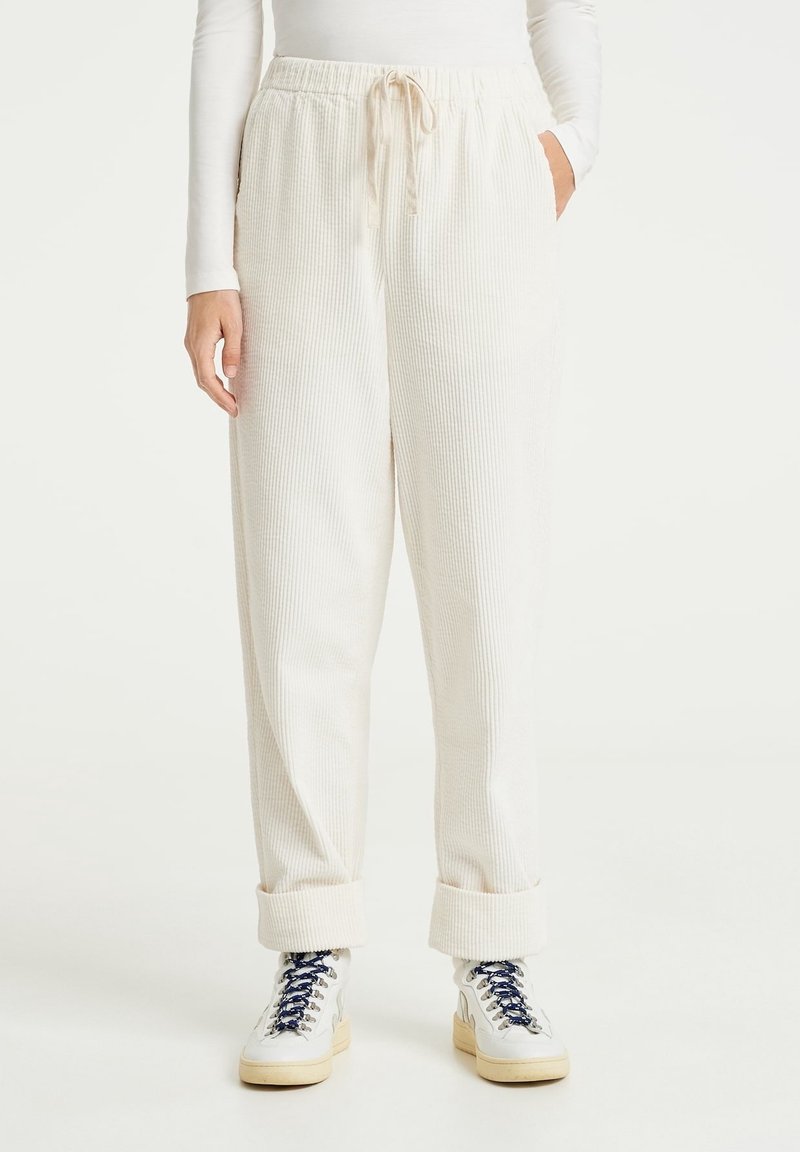 Personne portant un pantalon blanc côtelé avec taille élastique et cordon, revers remontés, associé à des baskets blanches avec des lacets foncés.