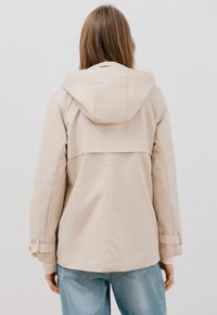 Beige huvjacka med avslappnad passform, texturerat tyg, vertikal söm bak och detaljer vid ärmarna. Bärs över ljusblå jeans.