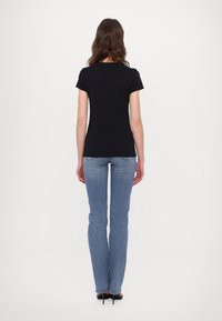 Tricou scurt negru din bumbac, purtat cu blugi drepti de culoare albastru deschis și pantofi cu toc negri, văzut din spate.