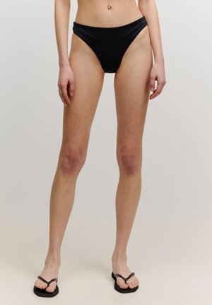 Schwarze Bikini-Höschen mit einem hoch geschnittenen Design. Glattes Material, minimalistischem Stil und einem schlichten, nahtlosen Bund. Kombiniert mit schwarzen Flip-Flops.