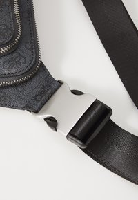 Matériau noir à motifs avec une surface texturée, boucle en métal argenté et sangle en tissu noir. Possède une poche zippée à l'avant.