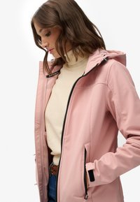 Chaqueta softshell de color rosa claro con capucha, con cremallera frontal, puños elásticos y bolsillos laterales. Se lleva sobre un jersey de cuello alto color crema.