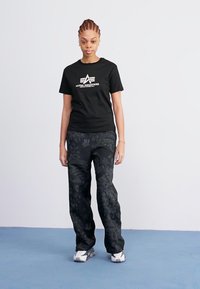 T-shirt noire avec le logo blanc "Alpha Industries", associé à un pantalon noir à motifs floraux. Le modèle porte des chaussures de sport grises et blanches.