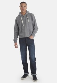 Grå hettegenser med kremfargede snorer, kombinert med mørkeblå denimjeans og svarte sneakers, brukt av en stående mannlig modell.