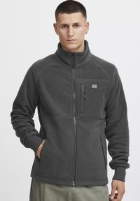 Adjustable gray fleece jacket with a high collar, zippered chest pocket, side pockets, and a smooth texture. Features a logo patch.
