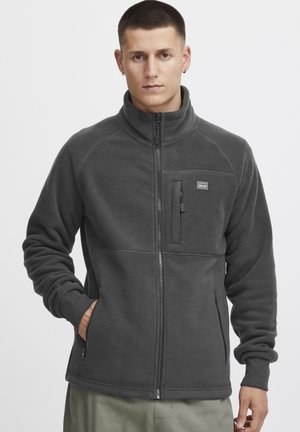 Adjustable gray fleece jacket with a high collar, zippered chest pocket, side pockets, and a smooth texture. Features a logo patch.