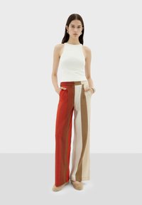 Top bianco senza maniche abbinato a pantaloni a gamba larga a vita alta con righe verticali rosse, marroni e beige. Tasche significative.