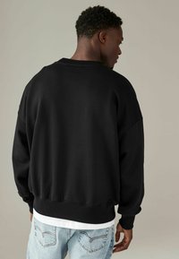 Zwarte oversized sweatshirt met een ribbelboord aan de hals en manchetten, met een zachte textuur en een relaxte pasvorm. Draagt over lichtblauwe jeans.