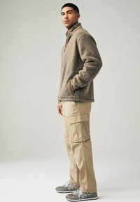 Fleece pulover svijetlosmeđe boje s patentnim zatvaračem i prednjim džepom; kombiniran s bež cargo hlačama koje imaju više džepova i opušteni kroj.