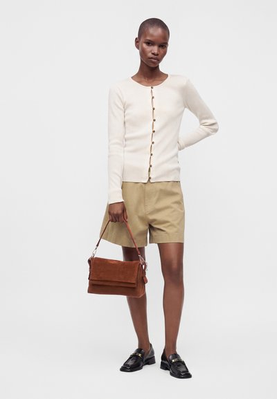 Femme portant un cardigan crème boutonné, un short beige, des mocassins noirs, tenant un sac à main en daim marron sur un fond clair uni.