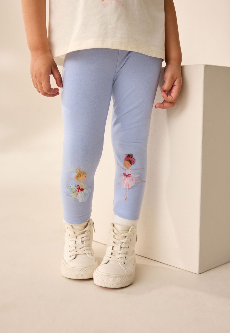 Kind trägt hellblaue Leggings mit Ballerina-Designs und weiße High-Top-Sneaker, steht neben einem weißen Block.