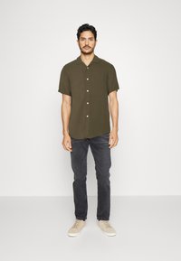 Chemise olive à manches courtes avec un col décontracté, devant à boutons et ourlet arrondi, associée à un jean foncé et des baskets de couleur claire.