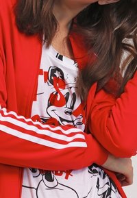 Červená zipová bunda s bílými pruhy, vrstvená přes bílý T-shirt s grafikou a textem černého a červeného Mickey Mouse.